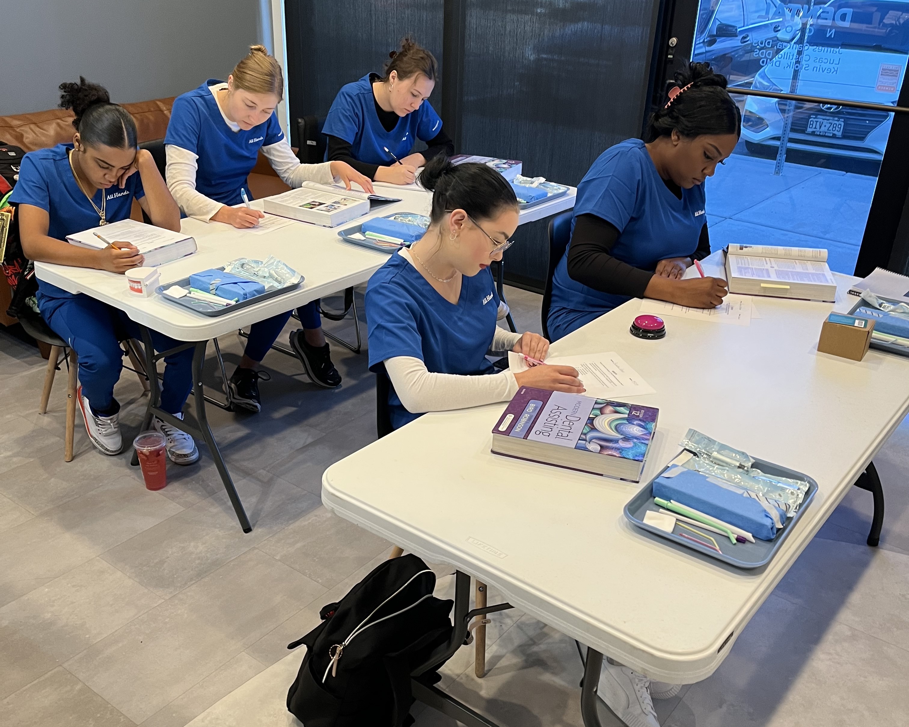 a group of people dressed in blue uniforms, sitting at white tables
            and studying on documents and books. They appear to be in a
            medical or healthcare environment, perhaps studying or training for
            some profession related to Dental Assisting at All hands dental
            school