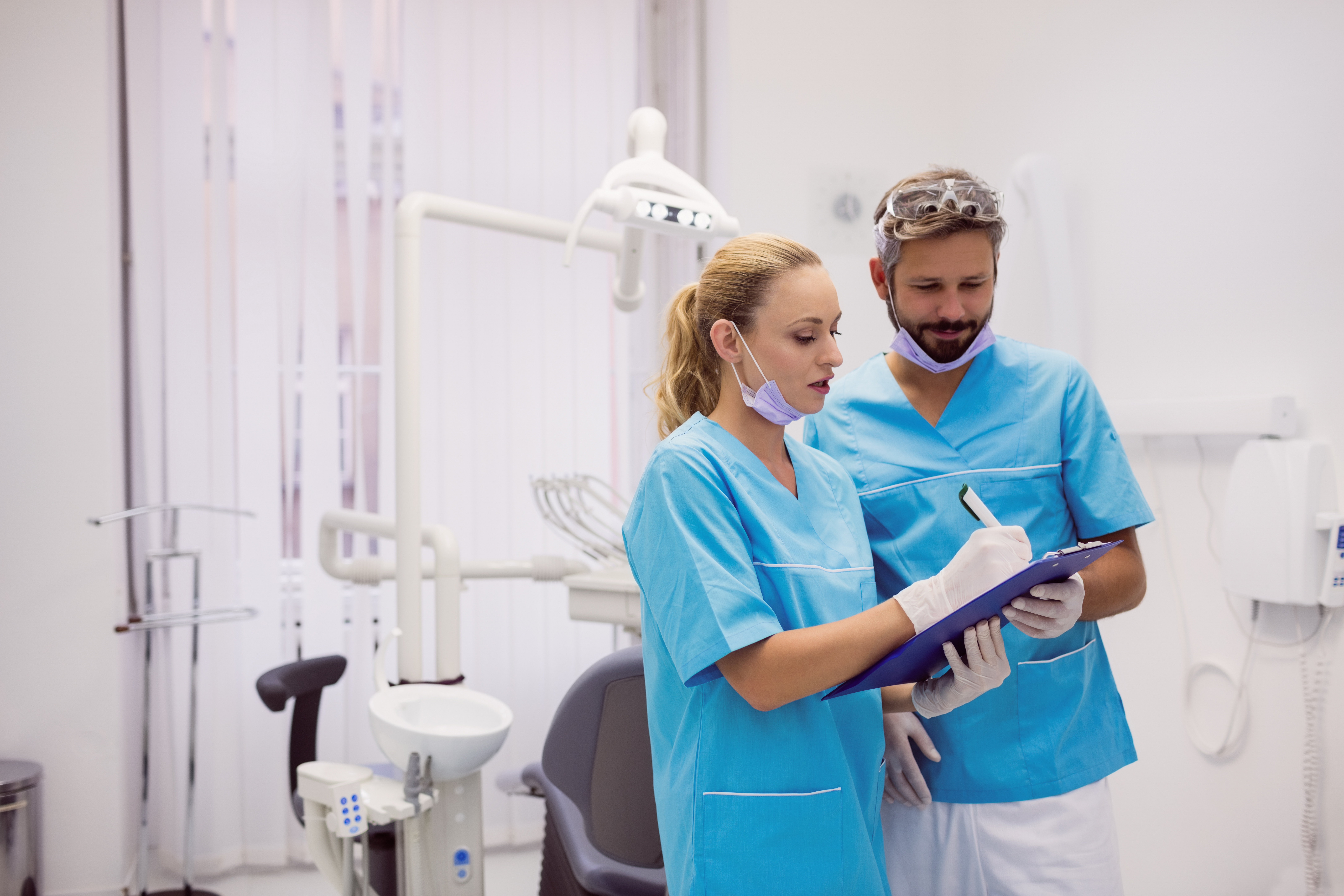 This image shows two people in blue medical uniforms in a dentist
                room. They are reviewing something on a clip board